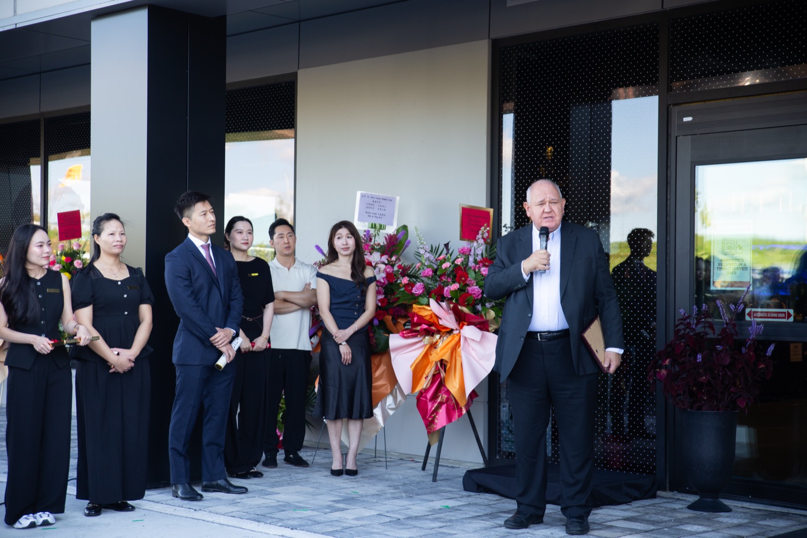 A guest speaking to the crowd outside the entrance, with staff and a floral arrangement alongside
